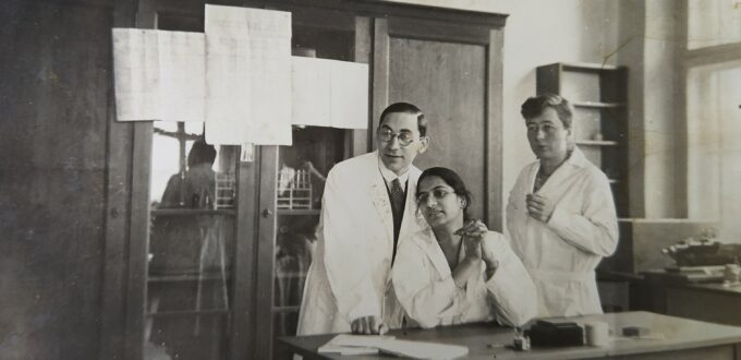 Irawati sits at a desk in the KWI-A’s doctoral students’ room. Two colleagues stand behind her. They all wear white coats and look interested to their right.